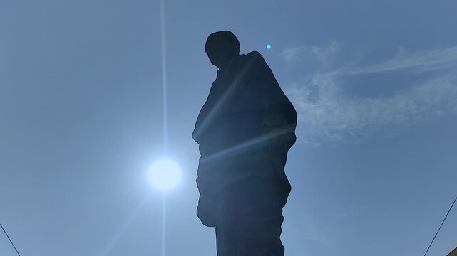 Extreme upward tilt of Statue of Unity in full silhouette with blazing sun and wide blue sky. Cinematic hero shot for India docs, patriotic content, and landmark title sequences.