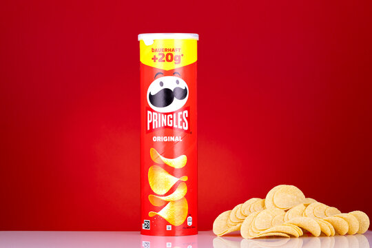 Illustrative editorial image of Pringles Original potato chips packaging with stacked crisps on a red studio background. Branded snack food product photographed in a clean commercial style.