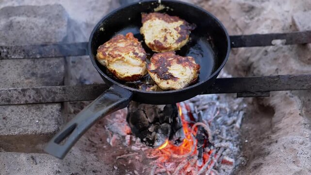 Juicy chicken steaks are fried in oil in a pan over an open fire until golden brown. The pan is on a makeshift stand. Around the hearth is sand. The fire is burning evenly. Smoke rises.