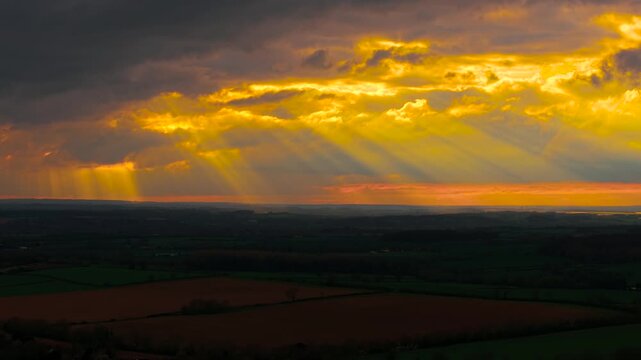 Ethereal sunlight shafts cutting through dense thundercloud layers across rural landscape at dusk. Fiery horizon illuminating fields under ominous sky formations. Moody cinematic atmosphere with