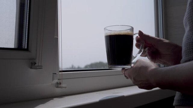 A tranquil morning scene unfolds as hands carefully place a steaming mug of black coffee onto a sun-drenched windowsill, capturing a peaceful and quiet moment of daily domestic life.