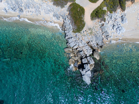 Aerial view of a pebble beach and rocky coastline with turquoise clear water and green vegetation in Pallini, Greece.