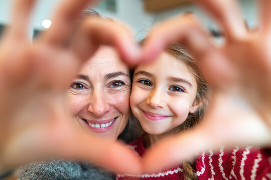 Mother and daughter form heart shape with hands, smiling together in a cozy indoor setting