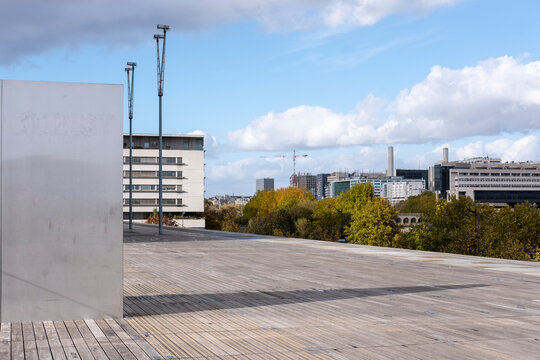 Open concrete terrace and plaza extend across urban architecture, with a building edge in exterior daylight beneath wide sky and distant trees
