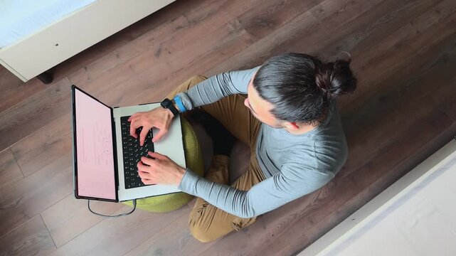 Unrecognizable Young Man Sitting on Floor and Typing on Laptop While Working Remotely at Home Viewed from Above in Bright Apartment Interior Authentic Freelance Lifestyle and Technology Scene