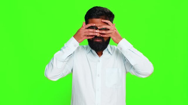 Indian man covers eyes with hands peeks through fingers, imitating a spy with dramatic expression of secrecy. Arabian guy isolated on green chroma key background showing mystery, tension hidden intent