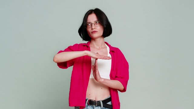 Young woman forms timeout symbol with her hands, looking serious to clearly indicate a request for a pause or break. Caucasian girl expresses the need to stop or slow down, isolated on gray background