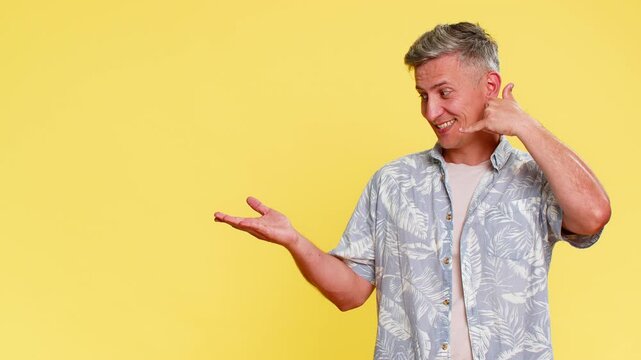Middle-aged man making call me gesture near ear, then confidently pointing to side, inviting to contact or call with clear intention. Guy expressing connection request isolated on yellow background.