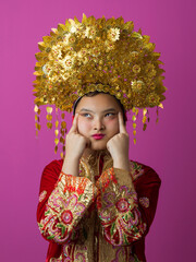 Fototapeta premium Young Indonesian Asian Girl Wearing Ornate Gold Headdress and Embroidered Traditional Costume Against Pink Background
