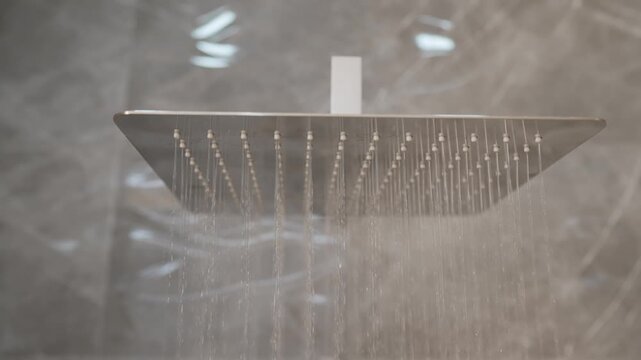 Close up of a modern square rain shower head. Water starts flowing from the nozzles in a contemporary bathroom with grey marble walls. Detail shot of running water