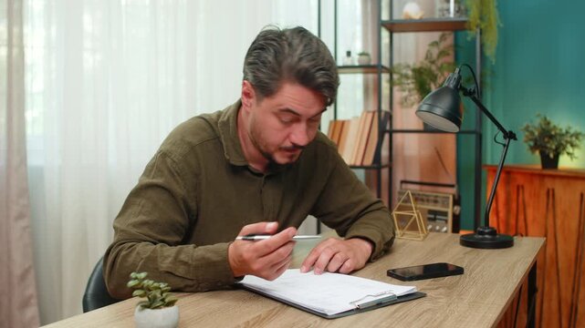 Middle-aged man at home reviews documents smiles satisfied signs papers approving content. Young adult guy at table proudly places signature feeling achievement confidence after final successful deal