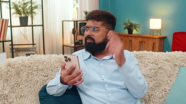 Indian man at home reads upsetting smartphone chat text brow knots shakes head shuts app feeling frustrated. Arabian guy on sofa makes no gesture shows disappointment mood rejects message negativity.