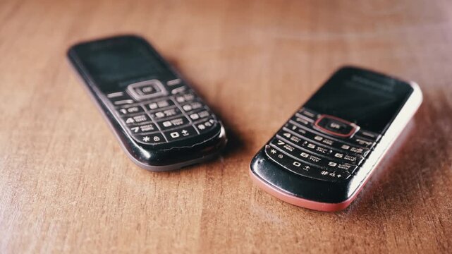 Two dusty vintage mobile phones with physical keypads sit side by side on a brown wooden surface. Concept of obsolete technology, electronic waste, and the evolution of digital communication