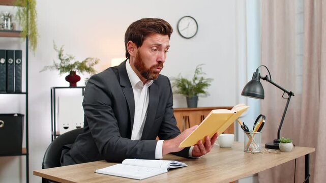 Middle-aged businessman at home office studies leadership guide smiles at quote and feels inspired to act. Freelancer guy at table writes idea in notebook sets clear goal and plans steps with care