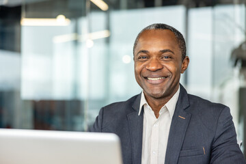 Fototapeta premium Confident mature executive smiling at camera, sitting at laptop in a modern office, portraying leadership, achievement, and positive corporate environment
