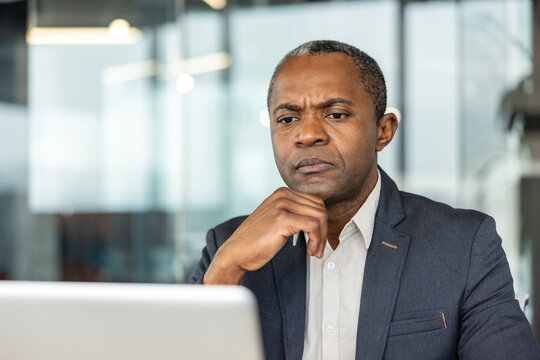 Serious businessman thinking deeply and frowning while viewing information on a laptop screen, conveying contemplation, problem-solving, and focused efforts in a corporate environment