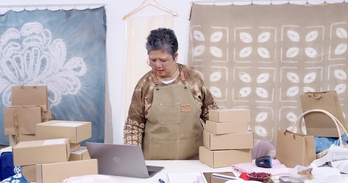 Senior artisan businesswoman smiles in slow motion, leans beside parcel stack, checks laptop order status, scans barcode label, lifts carton, prepares handmade textile shipping workflow home studio.