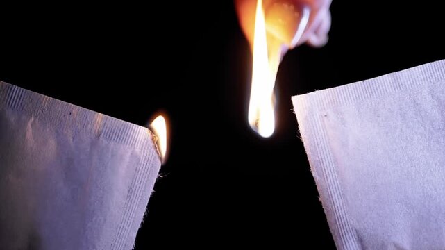 A hand uses a lit match to ignite torn edges of white paper packets against a black background. Concept of destroying evidence, starting fires, heat energy, and eliminating physical records.