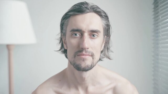 A serious young man with long hair and a beard gazing thoughtfully into the camera in a softly lit bedroom environment
