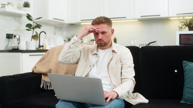 Young man at home looks at laptop sees defeat touches forehead sighs and clicks retry button. Caucasian guy on sofa watches screen shows frustration sad mood after bad news and failed attempt again.