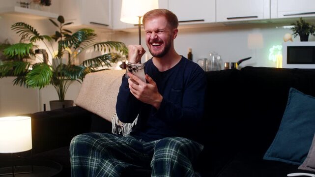 Young man at night home reads good news on smartphone smiles celebrates online game victory feels joy. Caucasian guy on sofa enjoys win raises fist happy reaction relaxes after work calm mood indoors.