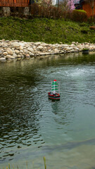 Obraz premium Small toy sailboat with green sails navigates a tranquil pond with a fountain