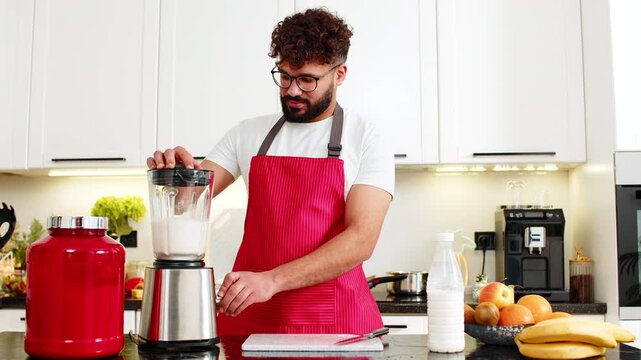 Arabian man blends milk whey protein banana apple smoothie for post workout recovery in kitchen at home. Young guy makes protein shake in apartment indoors feeling motivated after fit workout session.