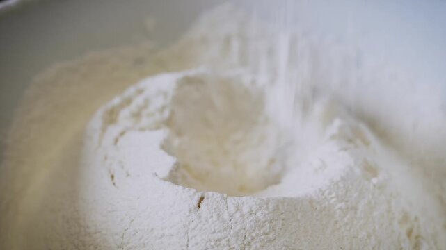 A close-up view of a white flour mound features a deep well in the center for mixing dough. Fine particles sift down to create a light texture. Soft lighting highlights the powdery.