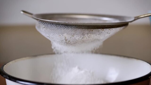 Metal kitchen sieve sifts white flour into a ceramic bowl in a slow motion close-up. Concept of homemade baking, culinary precision, artisan preparation, and healthy food.