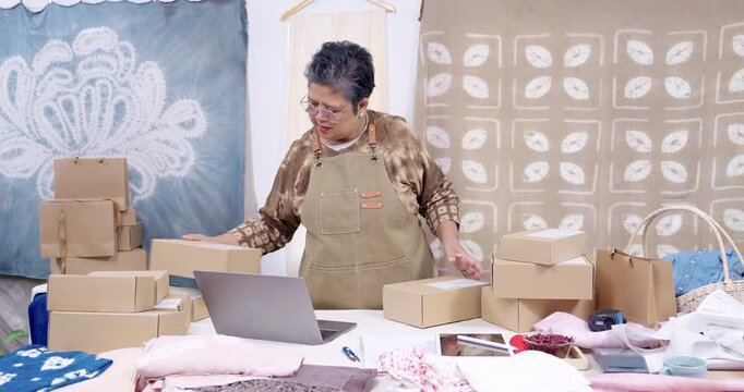 Senior asian businesswoman stands at home studio table, checks online order on laptop, arranges shipping boxes beside handmade shibori fabric, prepares packing workflow for parcel delivery today.