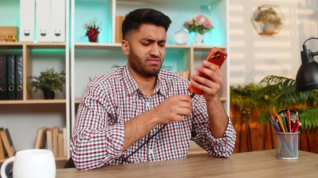 Indian man on home table checks broken charger cable and tries to charge smartphone with concern. Arabian guy on home table inspects phone problem and struggles with device battery during home work.