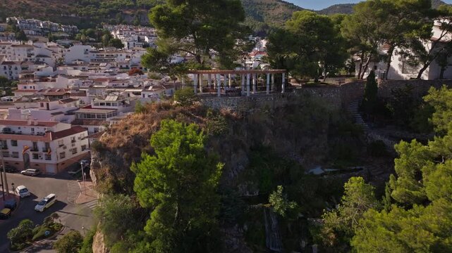 Drone footage pulling back and rising over Benalm&aacute;dena Pueblo's hilltop church, white arched facade, pine-covered knoll, parking areas, and terracotta rooftops backed by green mountains.