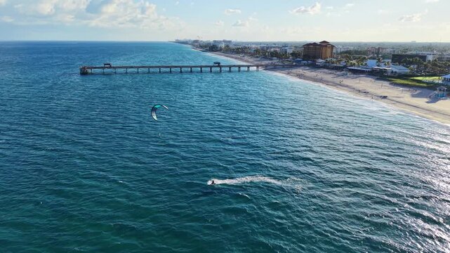 A kite boarder glides across the water near Deerfield Pier in Deerfield Beach Florida with the pier and coastline visible in the background