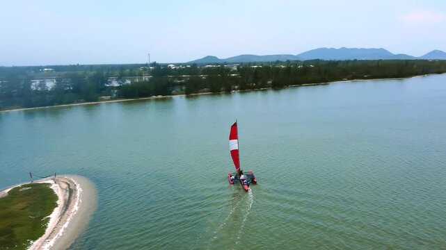 Trimaran navigating shallow lagoon water beside sandy shoreline in the Lakes Region of Rio de Janeiro State, Brazil, drone tracking shot
