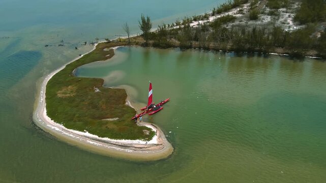 Trimaran rests beside a sandy island on Araruama Lagoon in the Lakes Region, Rio de Janeiro State, Brazil, drone orbit descent and push in above shallow hypersaline tropical lagoon water.
