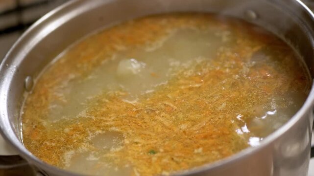 Grated carrots and broth simmer inside a stainless steel pot on a kitchen stovetop with steam rising from the surface. Concept of healthy eating, homemade nutrition, and culinary preparation