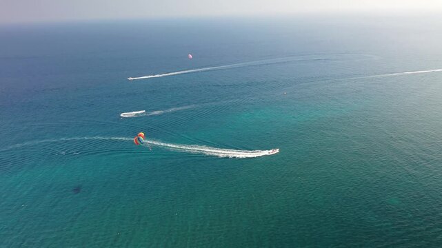 High-angle drone shot of speedboats cruising across the turquoise Mediterranean waters of Santorini. Highlights a colorful parasailer being towed, leaving white wakes on the open vibrant sea.