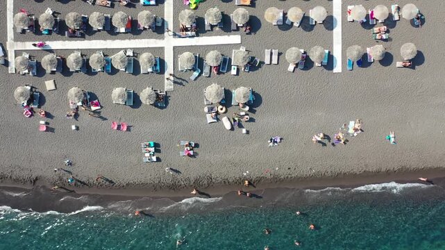 Wide drone shot showcasing the long black volcanic sand coastline of Kamari Beach. Features rows of umbrellas, coastal resorts, white-washed buildings, and the massive Mesa Vouno mountain cliff.