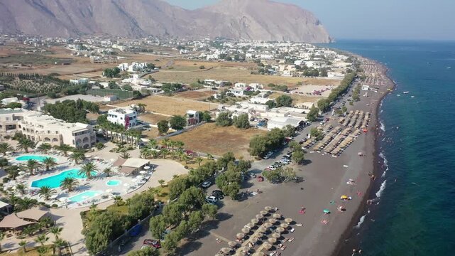 Wide drone shot showcasing the long black volcanic sand coastline of Kamari Beach. Features rows of umbrellas, coastal resorts, white-washed buildings, and the massive Mesa Vouno mountain cliff.