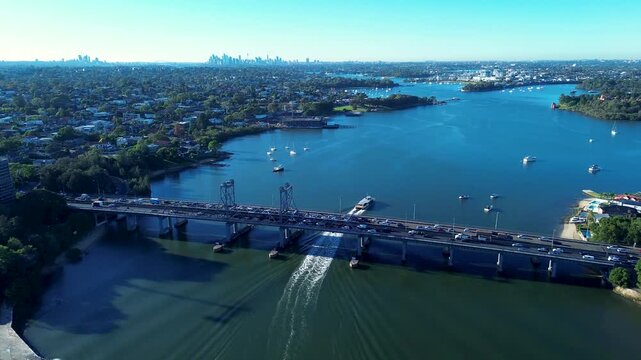 Drone aerial landscape of commuter ferry cruising along Parramatta River with car traffic traveling across Ryde Bridge and Sydney city skyline in the background near Meadowbank NSW Australia transport