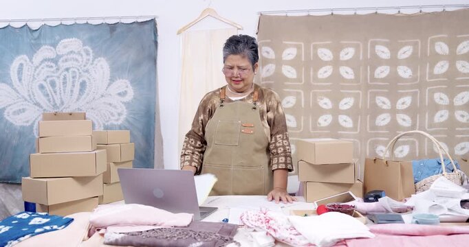 Senior asian entrepreneur reviews customer order paperwork, sorts invoice and receipt stack, checks parcel box count, updates online order status on laptop, prepares shipping batch in studio.