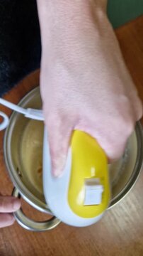 A hand holds a yellow electric mixer blending a smooth beige batter inside a stainless steel pot. The kitchen scene shows the mixing process from a top-down perspective on a warm wooden