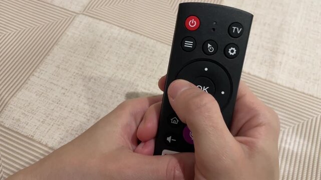 Remote control active. Close perspective of hands manipulating smart TV remote in living space. Intimate shot of hands engaging with streaming remote on decorative rug in cozy living environment