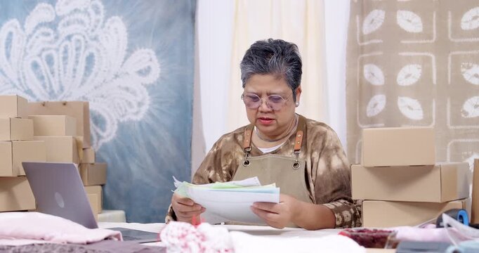 Senior asian businesswoman reviews accounting paperwork in slow motion, checks online order list on laptop, compares invoice with receipt, counts parcel boxes, manages shipping schedule in studio.