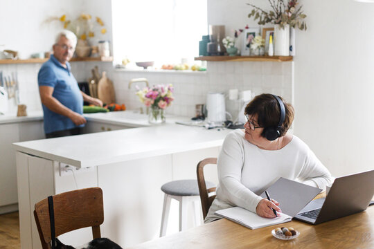 Portrait of senior woman working on laptop at wooden table in living room on background of modern kitchen and husband cooking. Mature business woman freelancer having work at home