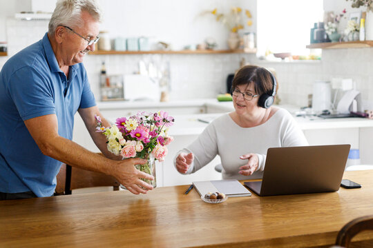 Senior husband giving flowers to working wife on laptop in living room. Domestic life and remote work. Happy Mothers day. Mature business woman receiving bouquet on holiday