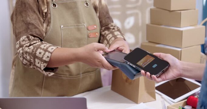 Slow motion payment scene, retired asian craft seller holds POS terminal, customer taps smartphone for NFC checkout, notes and tablet rest on table, parcels ready for shipping label printing.