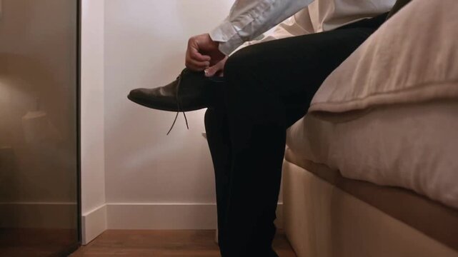 Close-up of a businessman sitting on a bed and putting on elegant black leather shoes, tying shoelaces in a modern bedroom