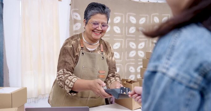 Older Asian female business owner hands POS terminal toward buyer, processes digital wallet transaction, maintains management desk, prepares handmade textile shipment for wholesale order logistics.