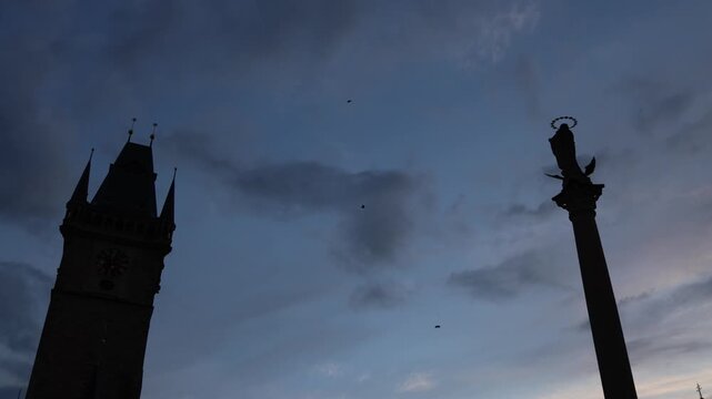 Paratroopers descending through dramatic evening sky above historic European square with dark silhouettes of old towers and monuments during aerial event